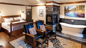 Ein Schlafzimmer mit einem hölzernen Bett, zwei Sesseln, einem Kamin aus Stein und einer Badewanne. An der Wand hängt ein Gemälde einer Flusslandschaft.