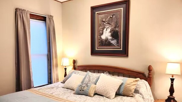 A bedroom with a bed, two bedside lamps, a framed picture on the wall, and a window with curtains. - Charming 4BR Craftsman Family Home in Emery (Emery)