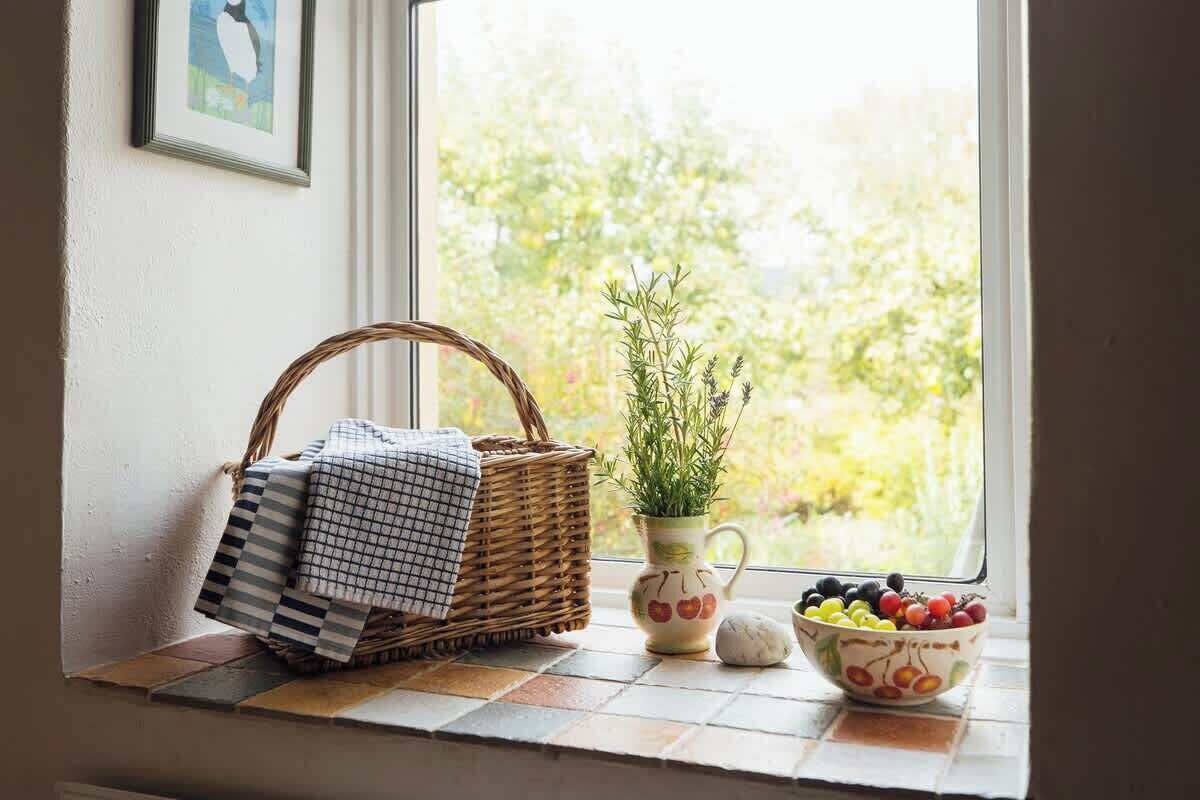 A window sill with a woven basket, a potted plant, a bowl of fruit, and a checkered cloth.