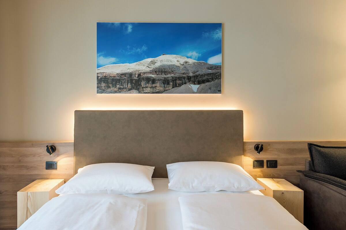 A hotel room with a bed, two bedside tables, a headboard, and a framed picture of a mountain landscape on the wall.