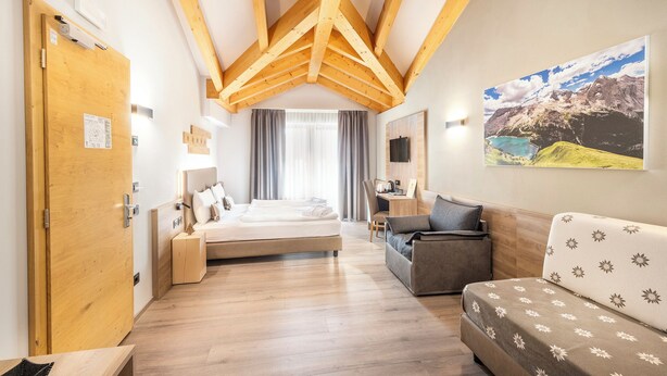 A modern hotel room with a wooden ceiling, a bed, a sofa, a desk, and a large window with a mountain view. - Chalet Aster (Moena)