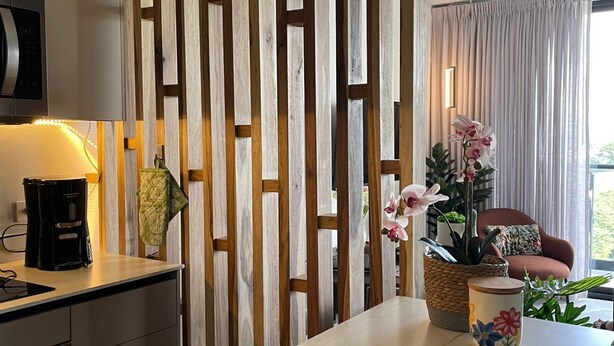 A modern kitchen with a wooden divider, a coffee maker, a fruit bowl, and a view of a living area with a sofa and plants. - Luxury Stay in Nucleo Sabana, AC, WIFI, Pools, Parking and 30 more amenities (San José)