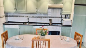 Une cuisine avec des meubles de rangement verts, une table à manger avec des chaises et un îlot central agrémenté d’un vase.