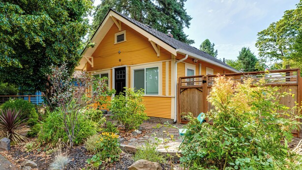 A yellow cabin with a wooden deck, surrounded by a garden with various plants and a rock formation.