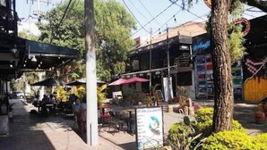 Un área con mesas y sillas al aire libre, rodeada de follaje y un edificio con un cartel.