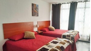 Habitación de hotel con dos camas, cada una con cabecero de madera, ropa de cama roja y una manta estampada. Hay un cuadro en la pared y una ventana con cortinas.