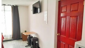 Una habitación con una puerta roja, un televisor montado en la pared, una mesita y una ventana con cortinas.