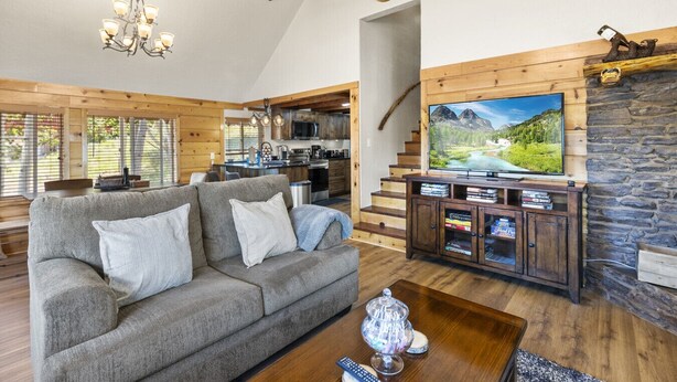 A cozy living room with a grey sofa, wooden beams, a stone fireplace, and a television displaying a scenic landscape.