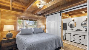 A bedroom with a wooden ceiling, a bed with gray bedding, a nightstand with a lamp, a dresser with a mirror, and a window with blinds.