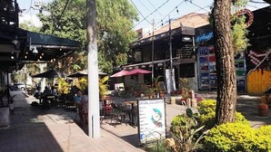 An outdoor café with tables and chairs, surrounded by greenery and buildings.