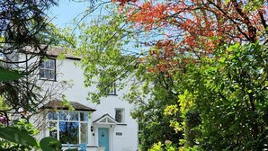 A gravel pathway leading to a white building with a blue door, surrounded by lush greenery and flowering plants. - Old Belfield, Bowness-on-Windermere with lake view (Bowness-on-Windermere)