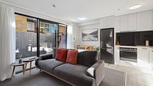 Un apartamento moderno con un sofá gris, una pequeña mesa de madera, una cocina con armarios blancos y un amplio ventanal con vistas a una pared de ladrillo y una terraza de madera.