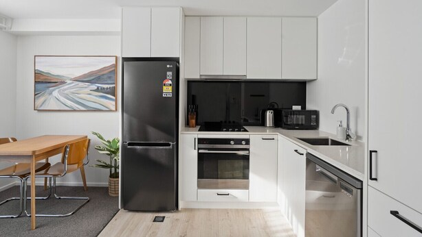 Una cocina moderna con armarios blancos, un refrigerador negro, un microondas, un fregadero y una zona de comedor con una mesa y sillas de madera.