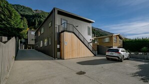 A modern building with a staircase leading to a wooden door, a white car parked in front, and a mountain backdrop. - Morepork Retreat - 1BDRM & Central (Queenstown)
