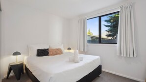 A bedroom with a large bed, two bedside tables with lamps, a window with curtains, and a view of trees and a building outside. - Morepork Retreat - 1BDRM & Central (Queenstown)