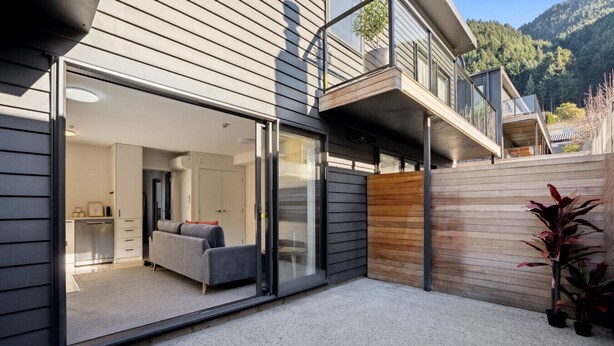 A modern house with a sliding glass door leading to a living area with a sofa and a dining table. - Morepork Retreat - 1BDRM & Central (Queenstown)