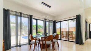A spacious dining area with a glass table and chairs, large windows with curtains, and a view of a swimming pool and outdoor seating. - Charming 3-bedroom villa in wonderful Ocean Village with Pool & BBQ (Sosúa - Caberete)