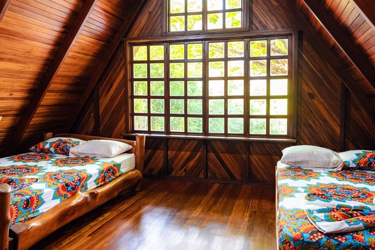 Quarto com teto de madeira inclinado, duas camas com roupa de cama colorida e uma janela grande com vários painéis.