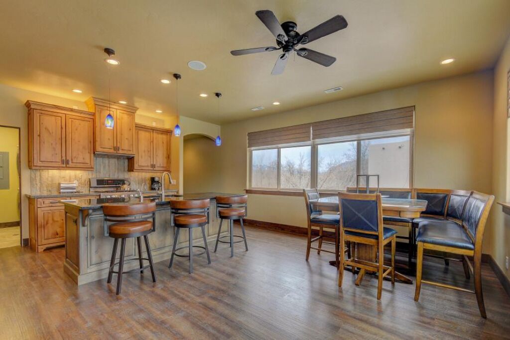 A modern kitchen with wooden cabinets, a central island with stools, a dining area with a table and chairs, and a ceiling fan.
