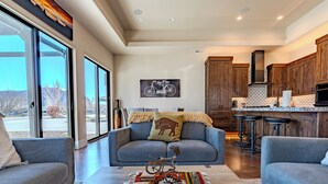 A modern living room with a gray sofa, a coffee table with decorative items, and a view of the kitchen and outside. - Village Camp Moab (Moab)