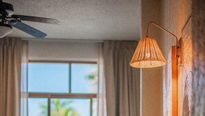 A bedroom with a bed, pillows, a wall-mounted lamp, a ceiling fan, and a window with a view of palm trees. - Pacífico Surf  (Puerto Escondido)