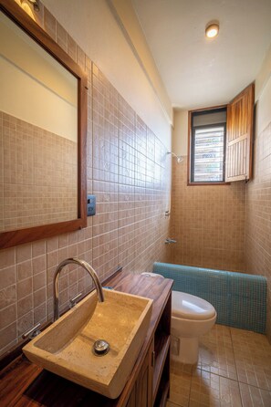 A bathroom with a beige sink, a mirror, a toilet, and a window with wooden shutters. - Pacífico Surf  (Puerto Escondido)