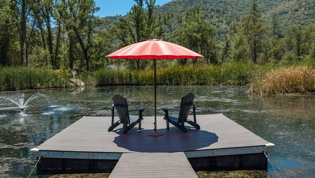 Un muelle de madera con dos sillas Adirondack y un paraguas rojo, con vista a un lago tranquilo y una ladera boscosa al fondo.
