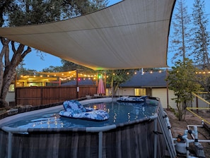 Une piscine extérieure avec des bouées gonflables, entourée d’une terrasse en bois et d’un espace couvert agrémenté de guirlandes lumineuses.
