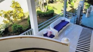 A balcony with two white sofas and blue cushions, a small table with a pink item, and a view of greenery and a building in the background.