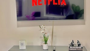 Uma TV de tela plana presa na parede, com a palavra "NETFLIX" em letras vermelhas. Abaixo da TV, há um centro de entretenimento preto com uma orquídea em vaso e duas figuras decorativas.