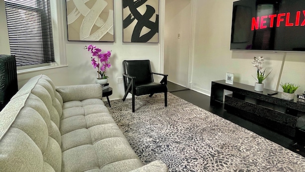 Uma sala de estar moderna com um sofá seccional, uma poltrona preta, um tapete estampado e uma TV de tela plana exibindo 'NETFLIX'.