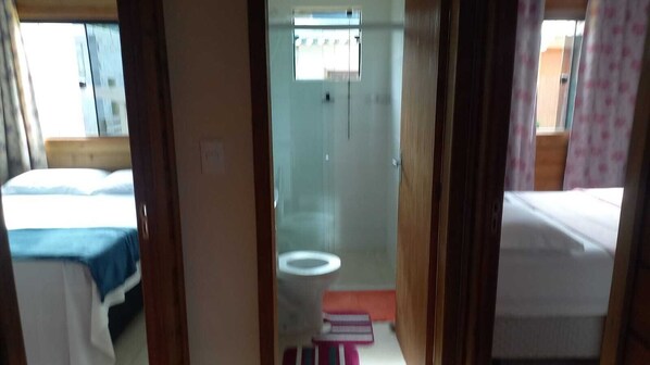 A narrow hallway leading to a bathroom with a toilet and a small window. There are beds with blue and white bedding on either side of the hallway.