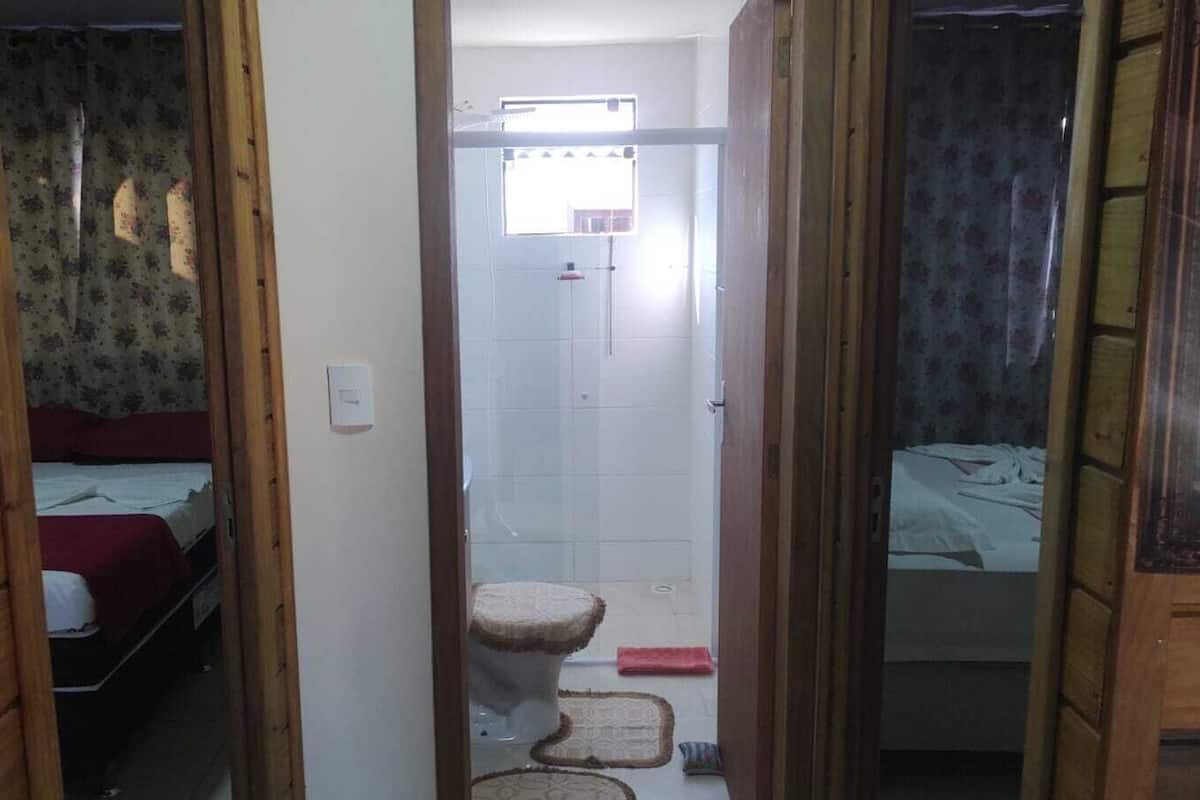 Un couloir étroit mène à une salle de bain équipée de toilettes, d’une douche et d’un lit recouvert d’une couverture rouge.