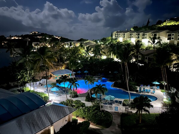A resort pool area with a clear blue pool, surrounded by palm trees and a white fence.