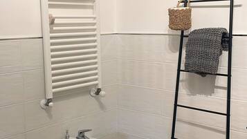 Une salle de bain comprenant des toilettes, un bidet, des murs recouverts de carreaux blancs, un plancher en bois, un porte-serviettes avec une serviette et un radiateur.