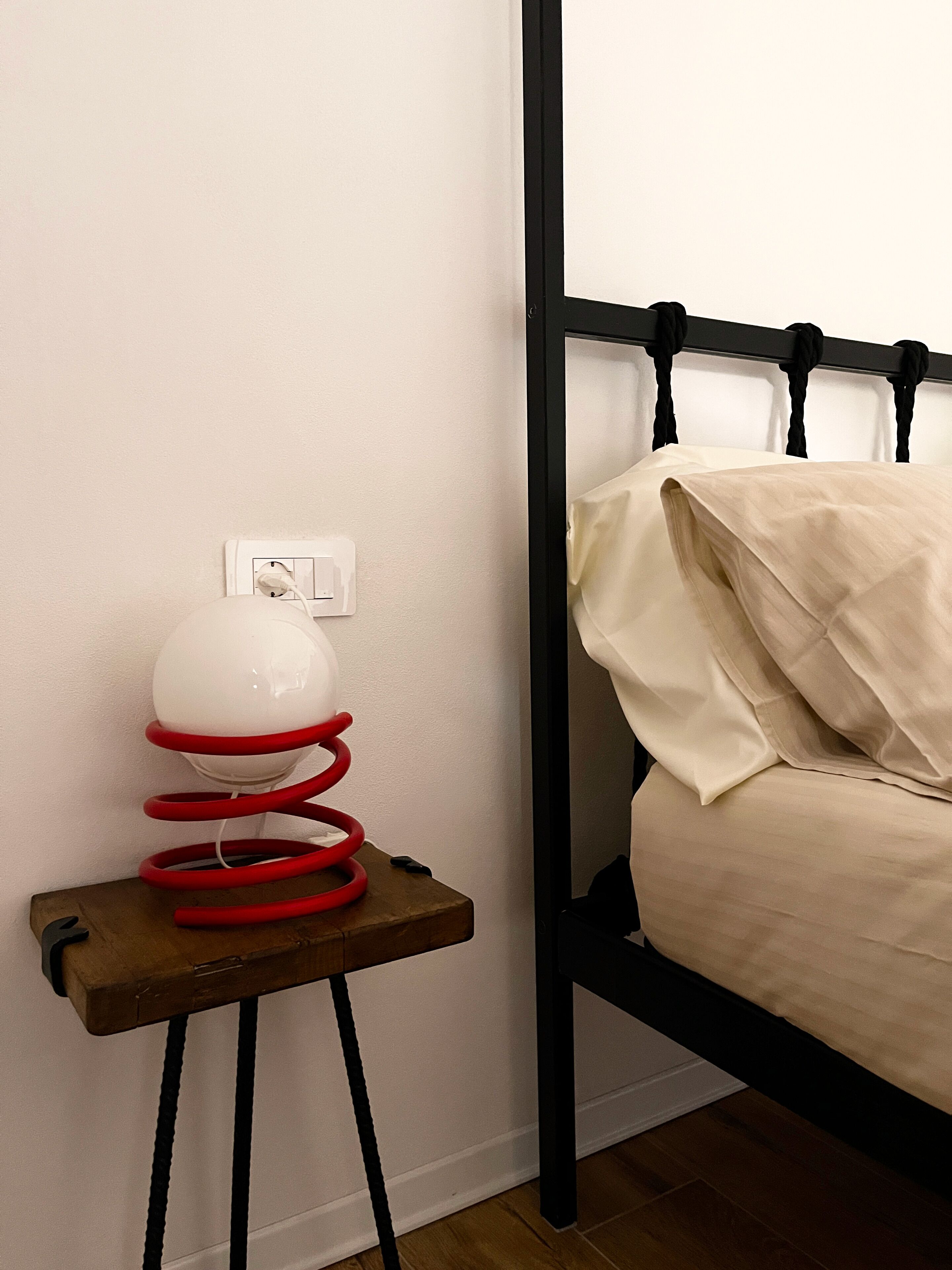 Un lit avec un cadre noir et du linge de lit blanc, une petite table de chevet en bois sur laquelle se trouvent un objet rouge enroulé et un objet blanc sphérique, et une prise électrique blanche sur le mur.
