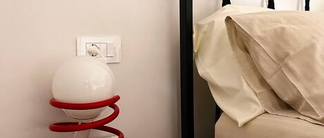 A bed with a black frame and white bedding, a small wooden nightstand with a red coiled object and a white spherical object, and a white electrical outlet on the wall.