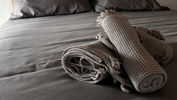 Un lit avec du linge de lit gris, deux oreillers et une couverture tissée.