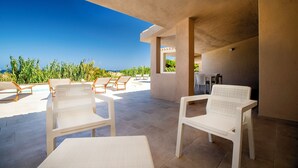 Zona de estar al aire libre iluminada por el sol, con sillas y una mesa blancas, con vista a una piscina y un cielo azul despejado.