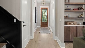 Un couloir doté d’un plancher en bois, de murs blancs, d’un escalier noir et d’une porte donnant sur une pièce avec une fenêtre.