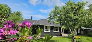 Ein schwarzes Holzhaus mit einem Ziegeldach, umgeben von üppigem Grün und blühenden Pflanzen.