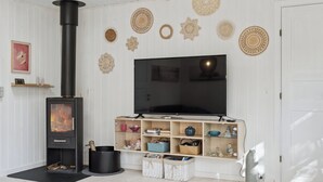 A living room with a black wood-burning stove, a flat-screen TV, shelves with decorative items, and a white door. - Charming cottage close to the beach (Glesborg)