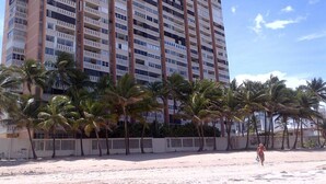 A high-rise hotel with multiple balconies, palm trees in front, and a sandy beach area.