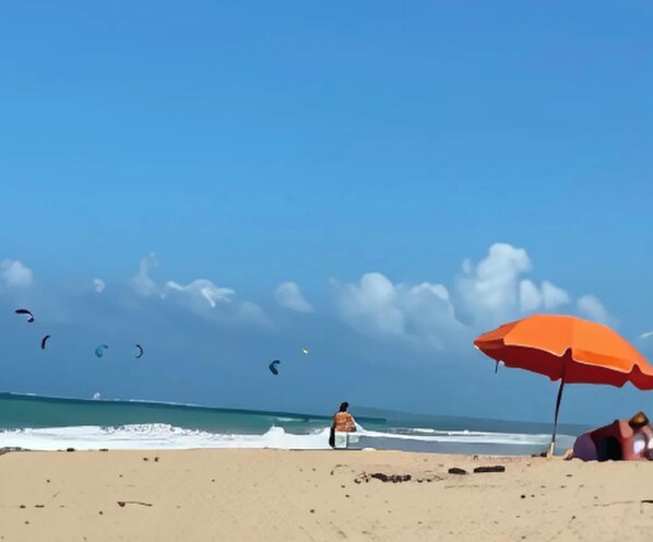Ein Strand mit blauem Himmel, einer Person im Schatten eines Sonnenschirms und fliegenden Drachen.