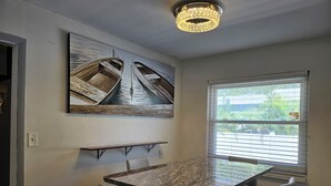 Un coin repas comprenant une table en marbre, quatre chaises, un lustre et un grand tableau représentant des bateaux accroché au mur.