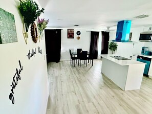 Cozinha moderna com ilha branca, área de refeições com mesa e cadeiras pretas e sala de estar com cortina preta.