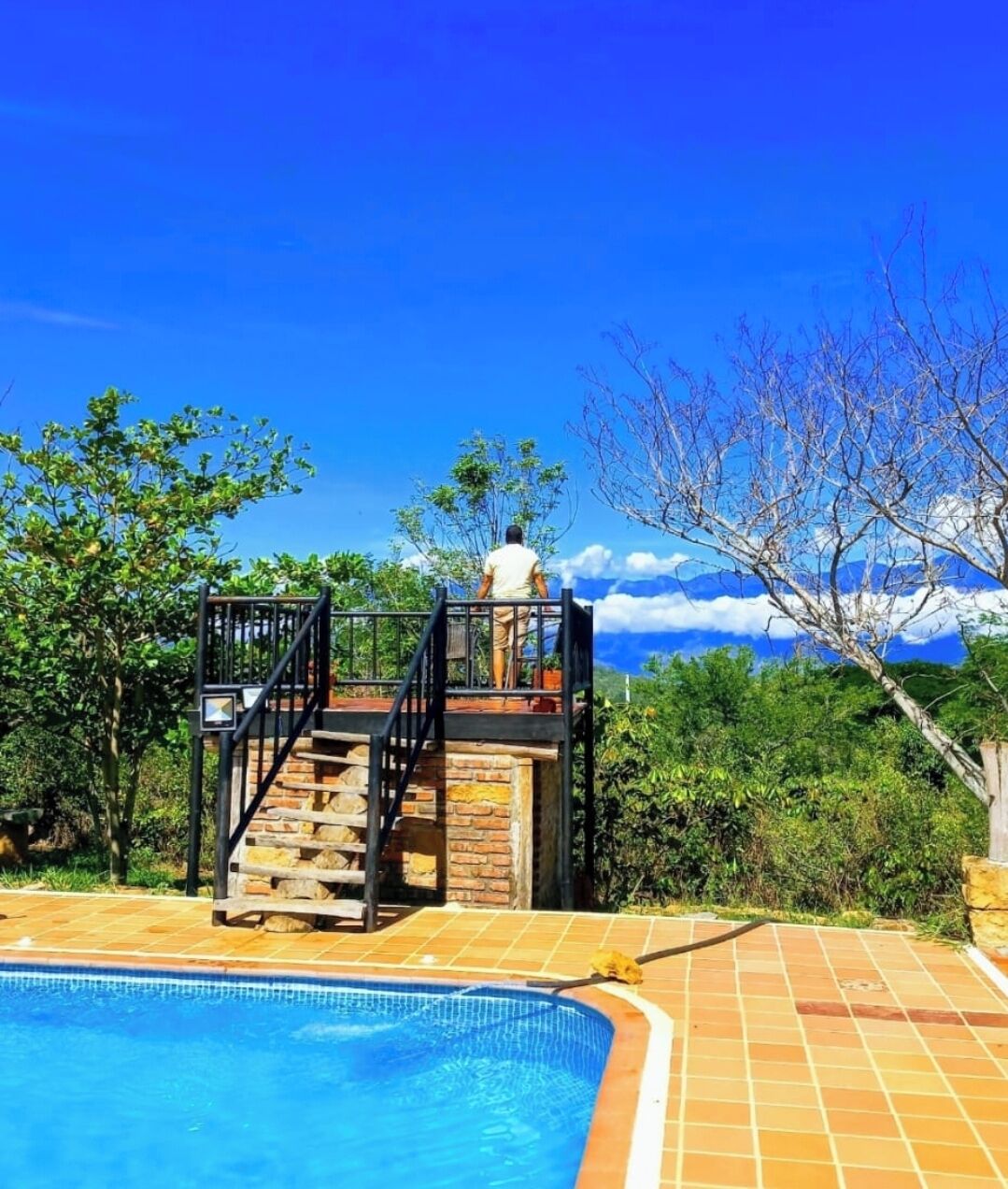 Une piscine avec un escalier en pierre menant à une plateforme d’observation, entourée d’une végétation luxuriante et surplombant un ciel bleu azur.