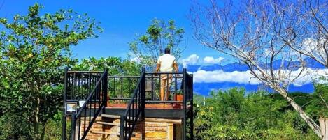 Une piscine avec un escalier en pierre menant à une plateforme d’observation, entourée d’une végétation luxuriante et surplombant un ciel bleu azur.