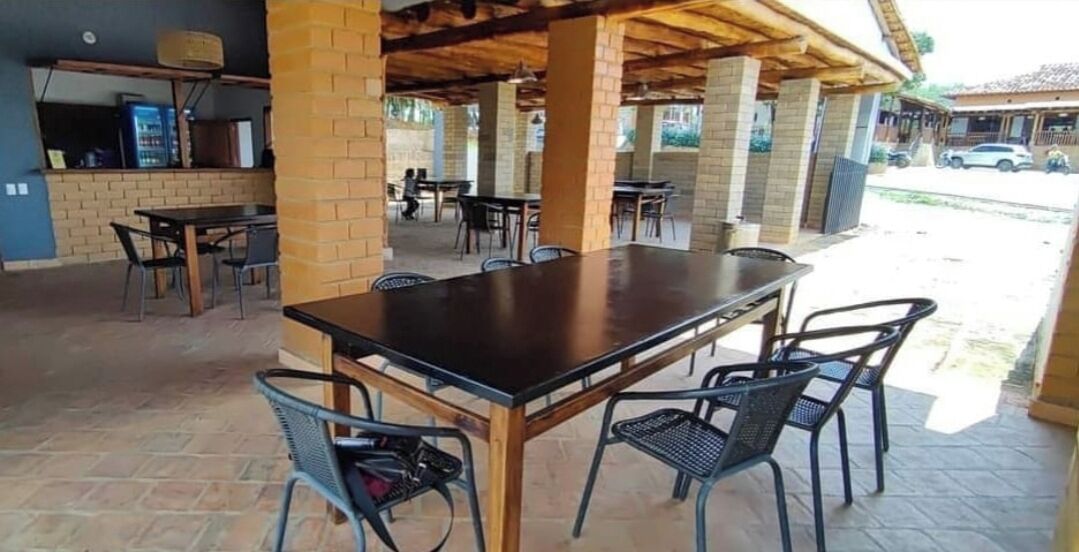 An outdoor patio with a long wooden table and metal chairs, brick pillars, and a covered area with a brick wall and a window.