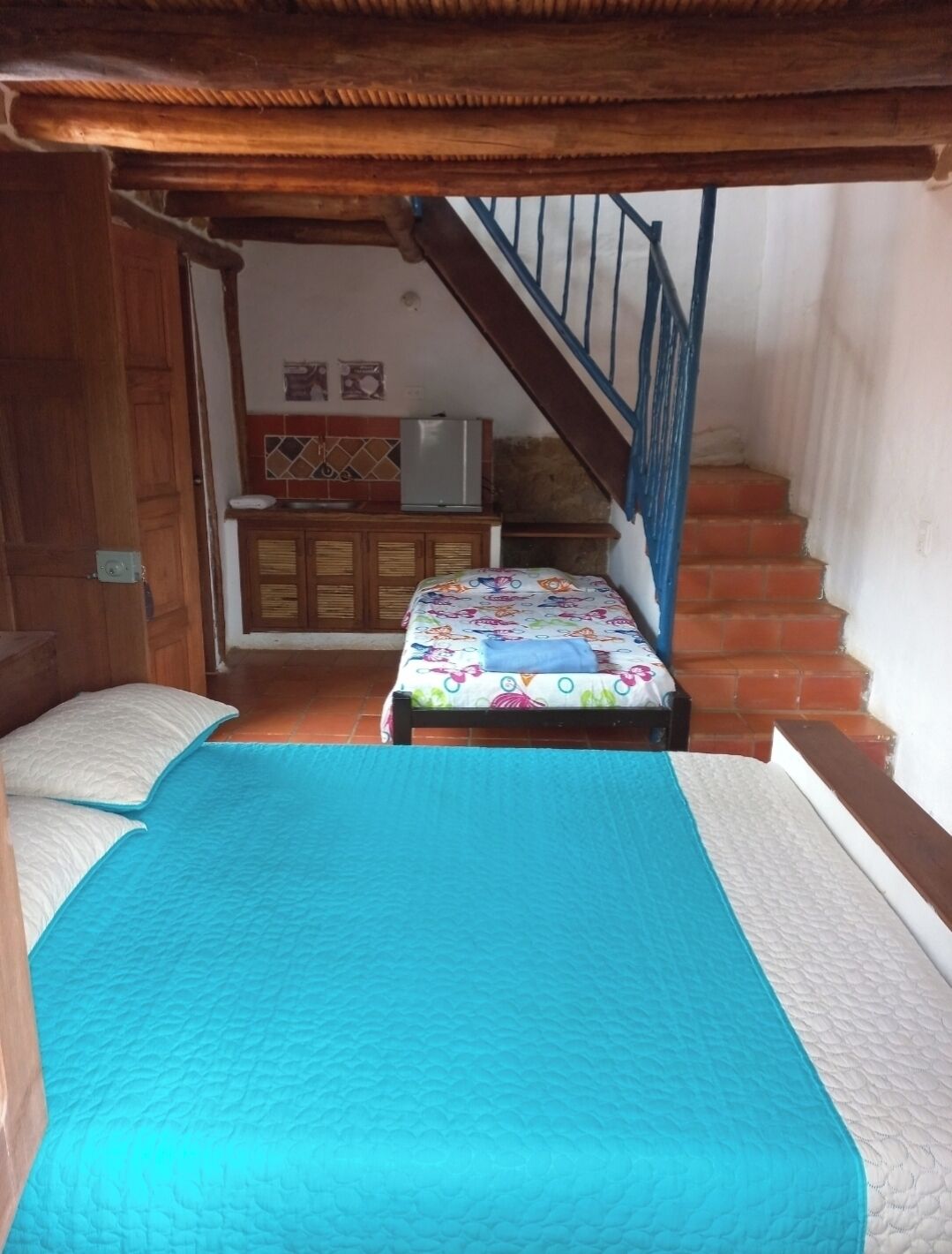 A bedroom with two beds, a wooden ceiling, a staircase, and a kitchen area in the background.